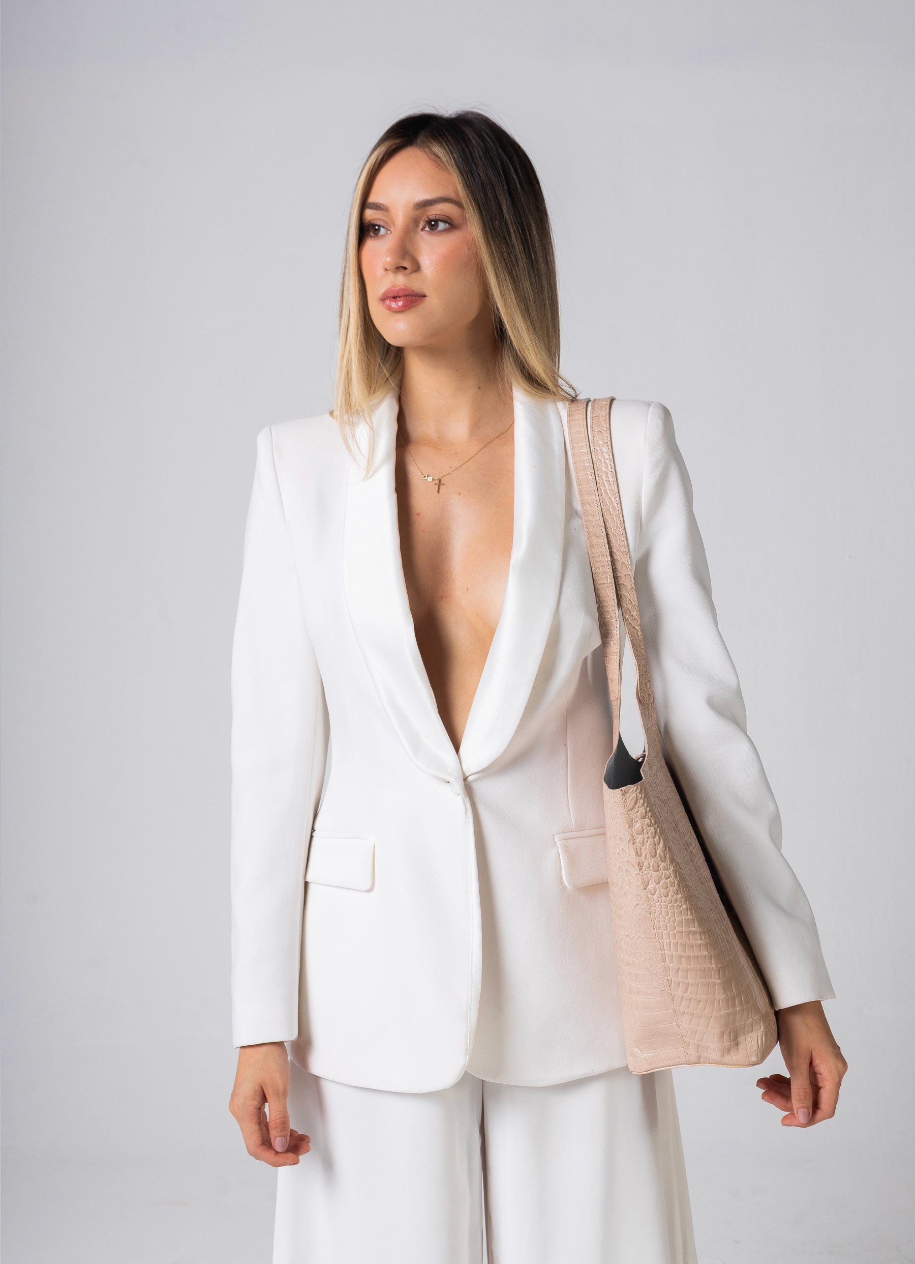 A woman modeling a white tailored blazer and matching trousers, styled with a beige woven shoulder bag against a light gray studio background.