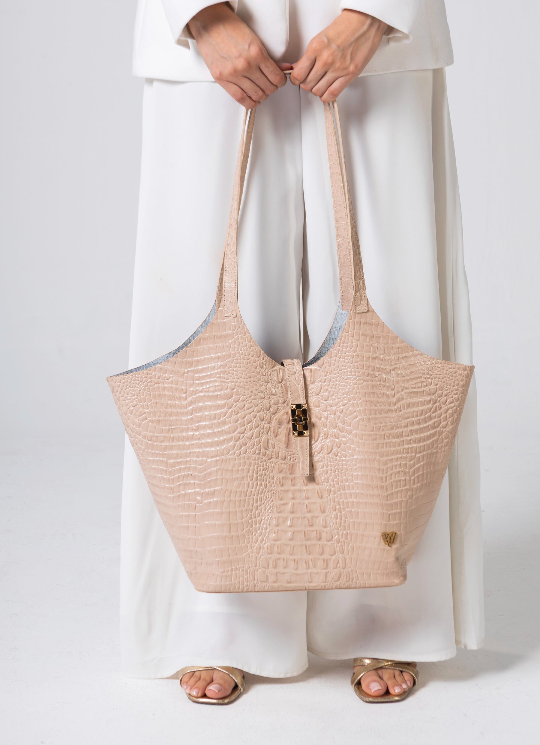A person in a white outfit holding a large beige crocodile-embossed tote bag with long shoulder straps and a small gold heart logo, photographed against a light gray studio background.