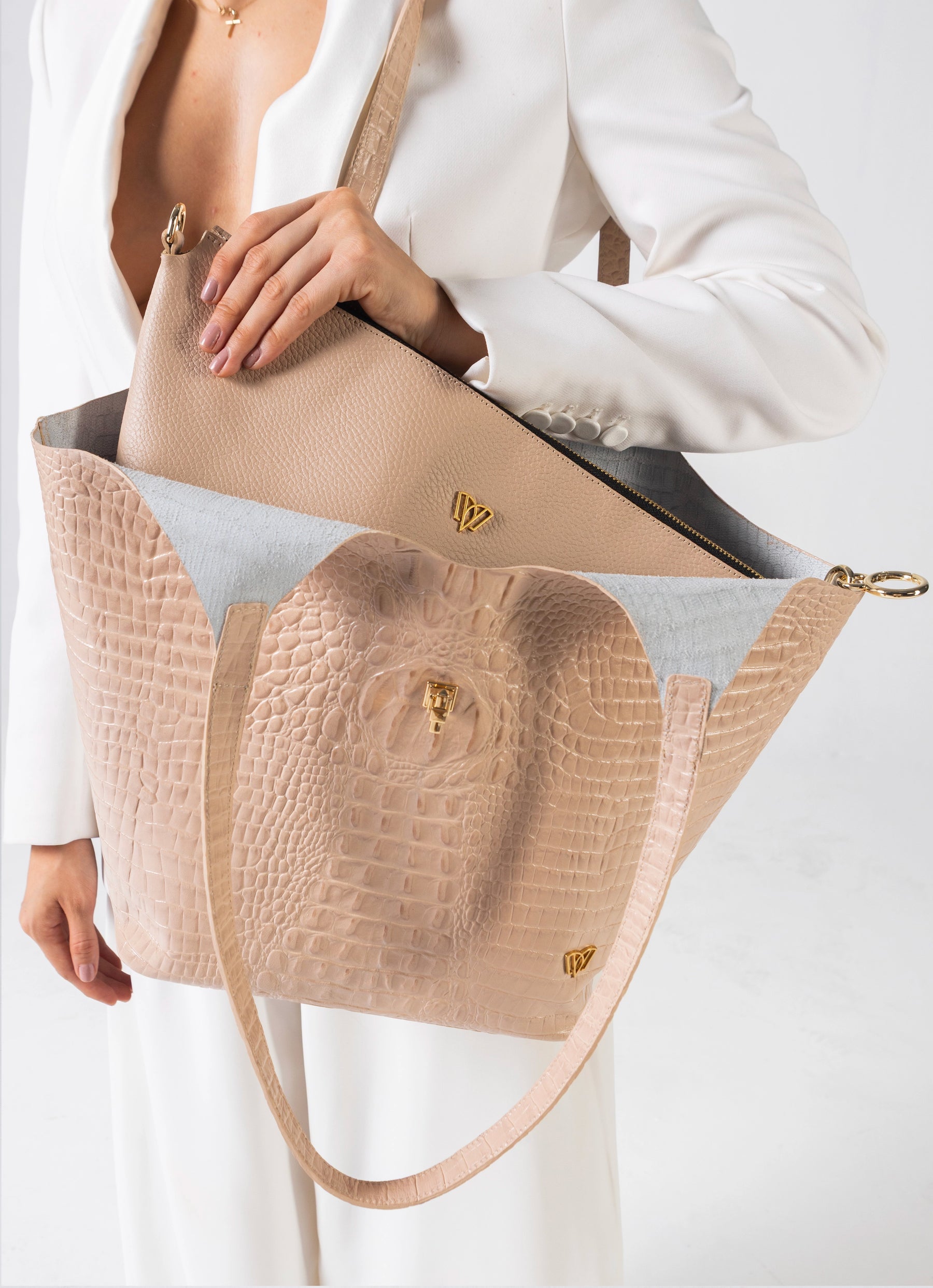 A beige tote bag with a crocodile-embossed front panel, a smooth beige flap, and gold-tone hardware, shown being held by a model wearing a white blazer.