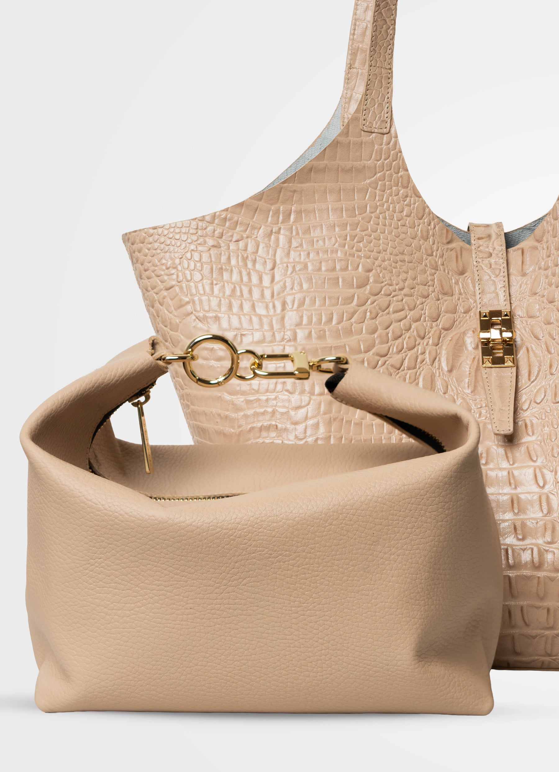 A beige pebbled leather shoulder bag in the foreground with gold-tone hardware, positioned in front of a larger beige crocodile-embossed tote bag on a light gray studio background.